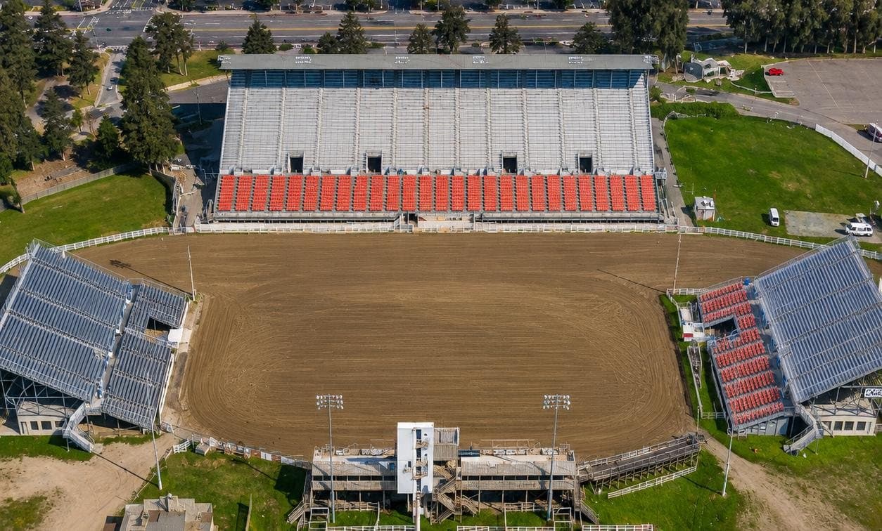 Overhead view of the rodeo arena at the Salinas Sports Complex, showing the main covered grandstand with rows of orange seating along one side of the dirt arena and two further grandstands angled along the sides.